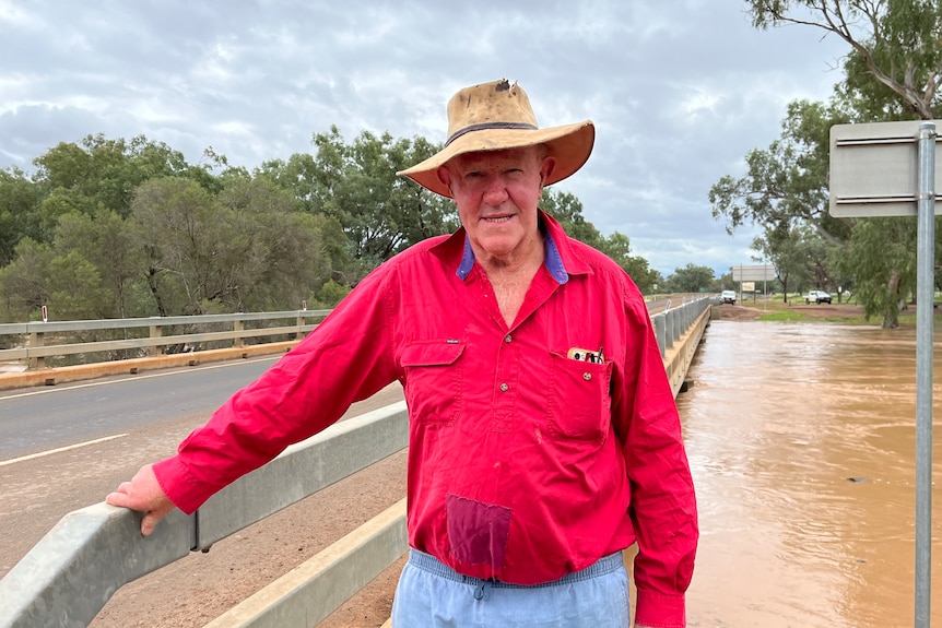 a farmer in a red shirt and akubra standing on a bridge above a flooded river