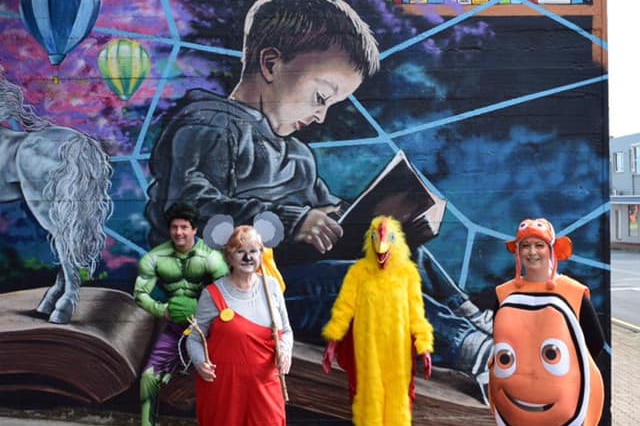 Library staff dressed up in book week costumes stand in front of a mural.