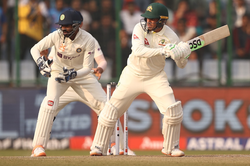 An Australian male batter watches a shot to the leg side as the Indian wicketkeeper looks on.