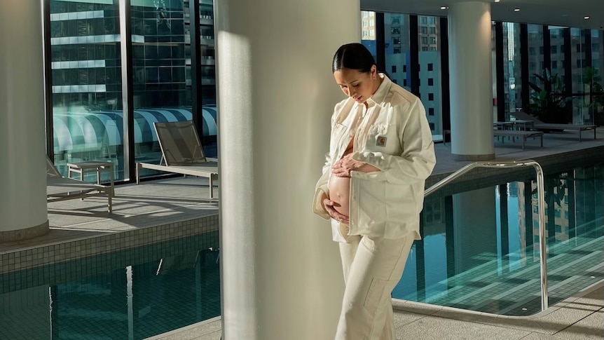 A picture of a woman in white pants and open shirt cradling her pregnant belly while standing by an indoor pool.