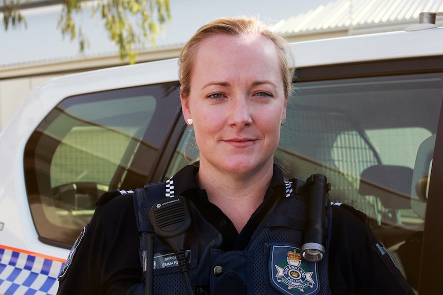 A woman in police uniform looks straight at the camera, standing beside a police 4WD vehicle.