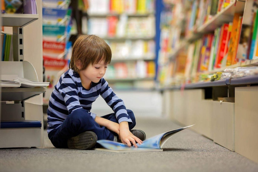 A young boy sits on the floor of a boy store reading a book that rests on the ground.