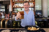 A man behind a bar holding a beer up to the camera and smiling
