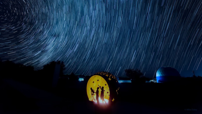 A silhouette of two people standing next to a telescope looking up at a starry night sky. 