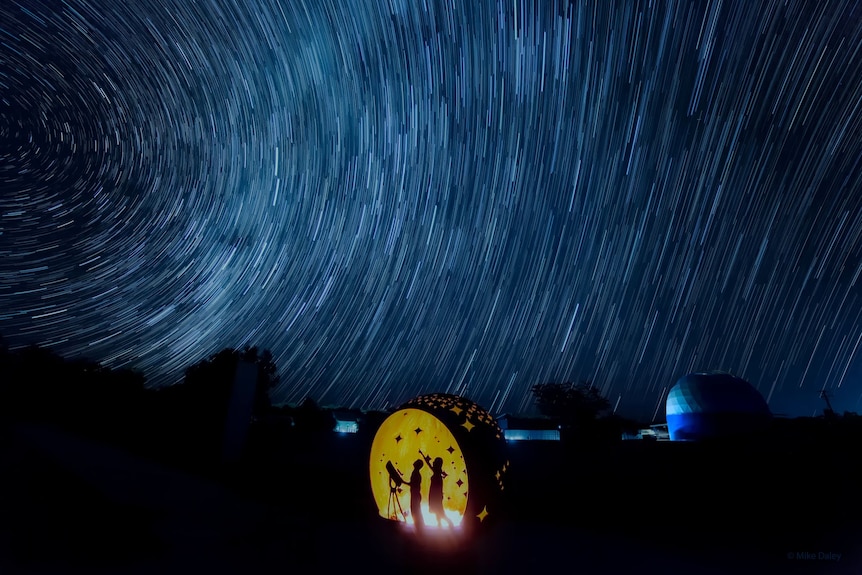 A silhouette of two people standing next to a telescope looking up at a starry night sky. 