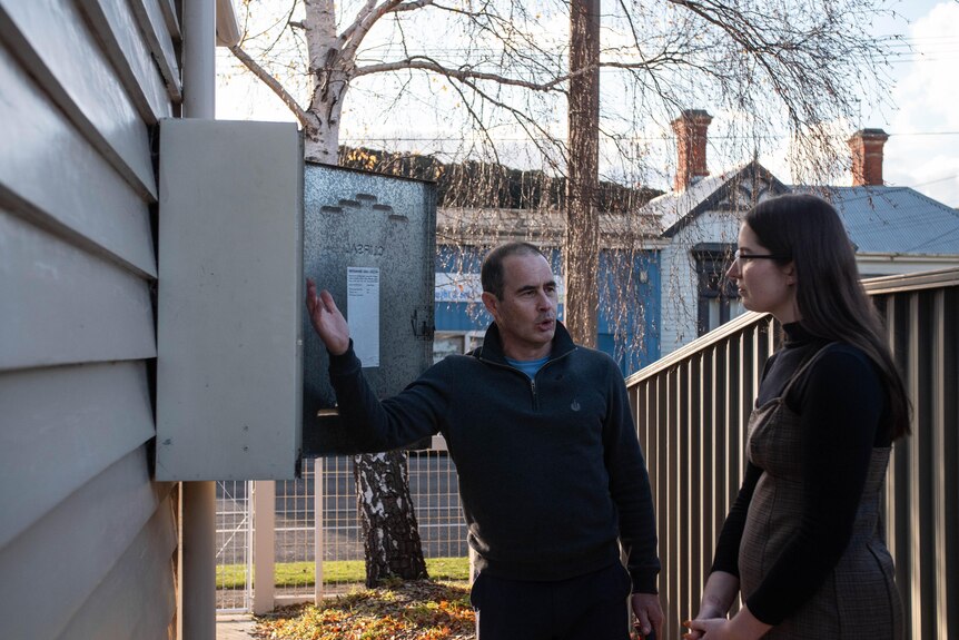 A man gestures toward an open fuse box on the side of a house, as he speaks to a woman