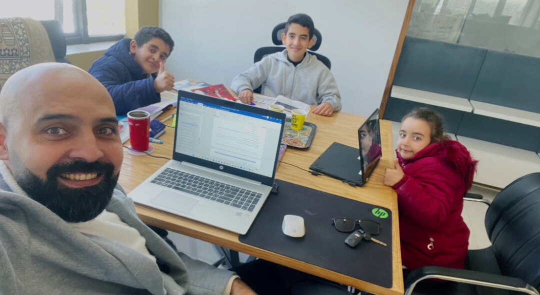 Man sitting at a desk with his three children, and a laptop in front of him.