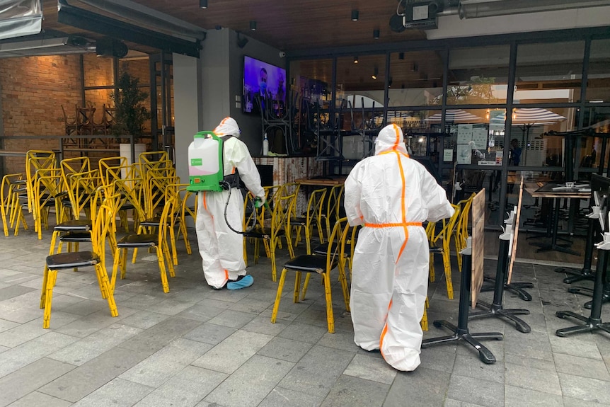 Two people in protective gear clean tables and chairs outside a restaurant.