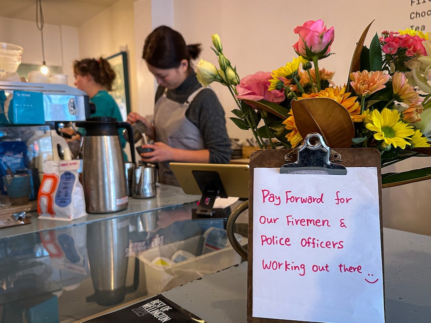 A sign on a cafe bench reads "Pay forward for our firemen and police officers working out there" next to a bunch of flowers