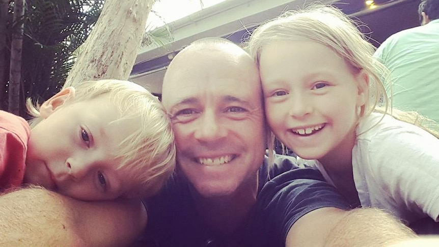 A selfie of a father with a young boy resting his head on one shoulder and a young girl smiling over the other shoulder.