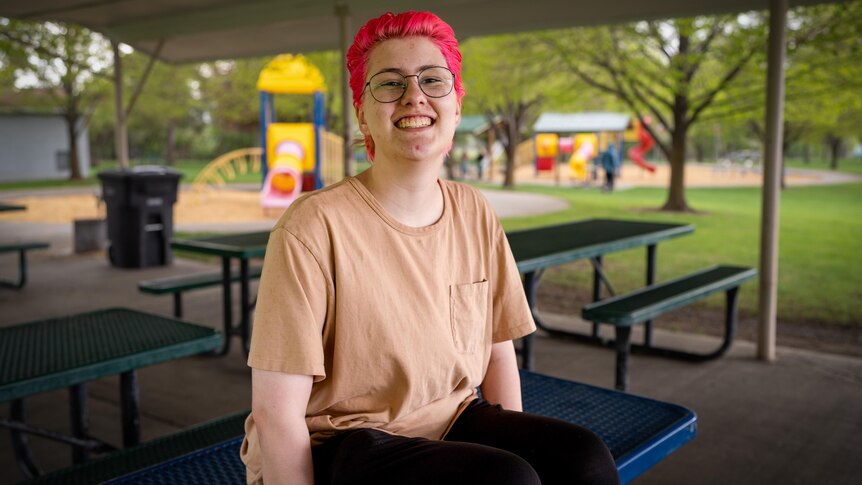 A teenager with pink hair sits on a park bench, smiling 