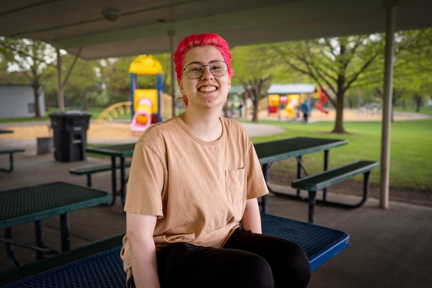 A teenager with pink hair sits on a park bench, smiling 