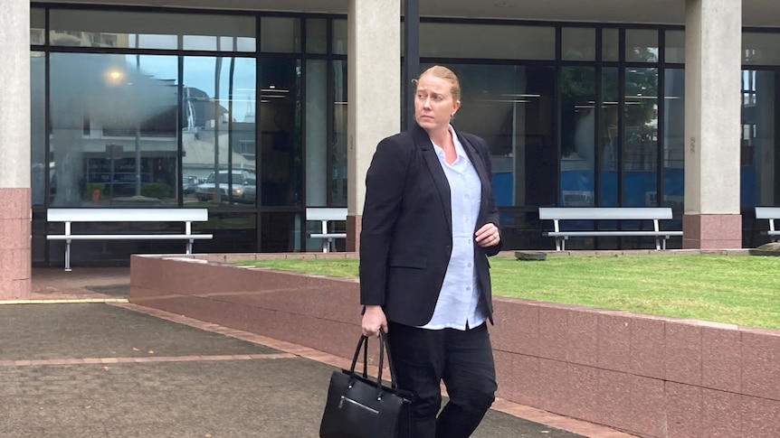 A woman wearing a black jacket and a white shirt, holding a black handbag, walks outside a building.