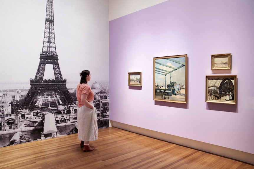 A woman standing in a gallery looking at four paintings on a wall with a large image of the Eiffel Tower behind her.