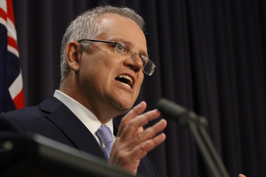 Side view of Scott Morrison speaking against a dark backdrop, hands gesturing, looking serious.