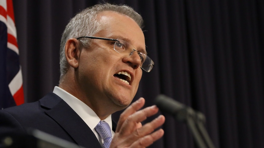 Side view of Scott Morrison speaking against a dark backdrop, hands gesturing, looking serious.