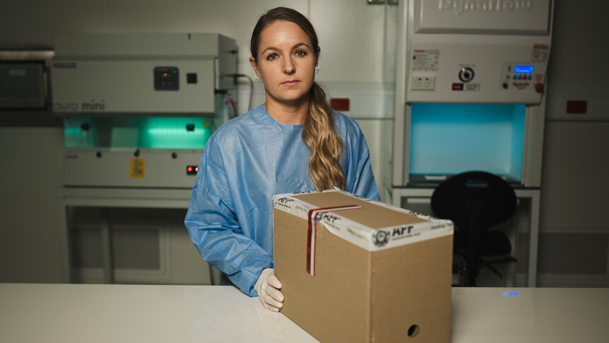 Woman in PPE holding a cardboard box on a table.