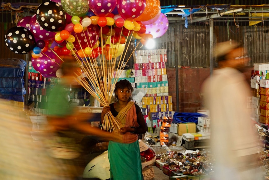 A young girl holding a bunch of balloons walks down a busy street at night.