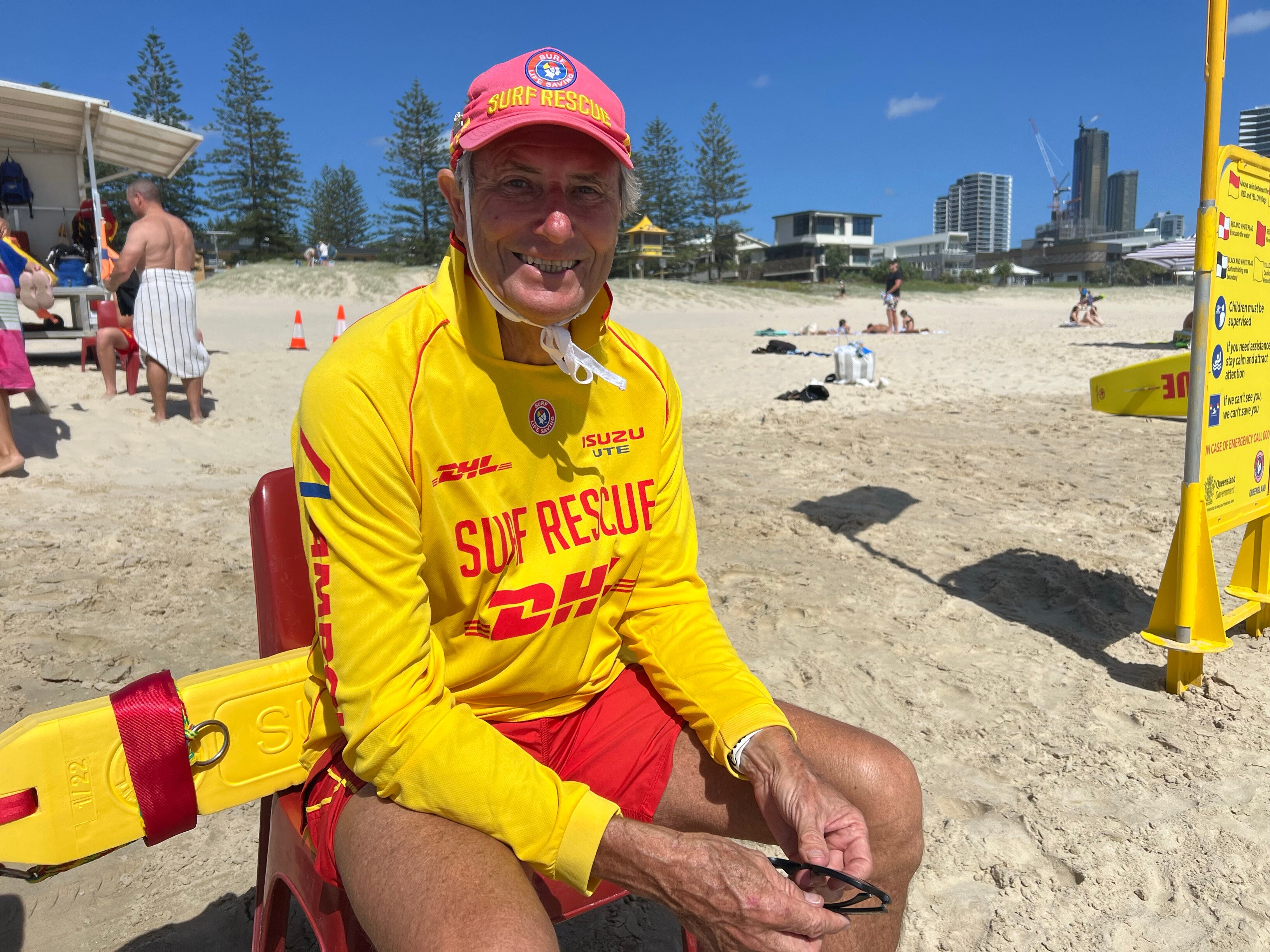 Man in yellow and red lifesaving outfit sits on beach and smiles