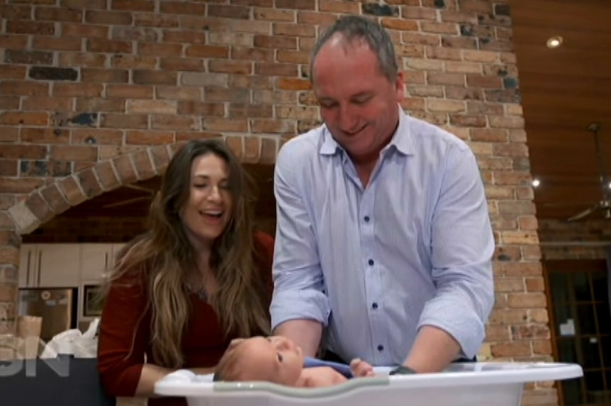 Barnaby Joyce holds his infant son in a baby bath while Vikki Campion looks on, smiling.