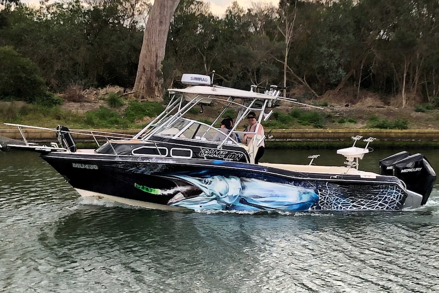 A blue boat is in the water, it has a fish painted on the side and three people on the boat.