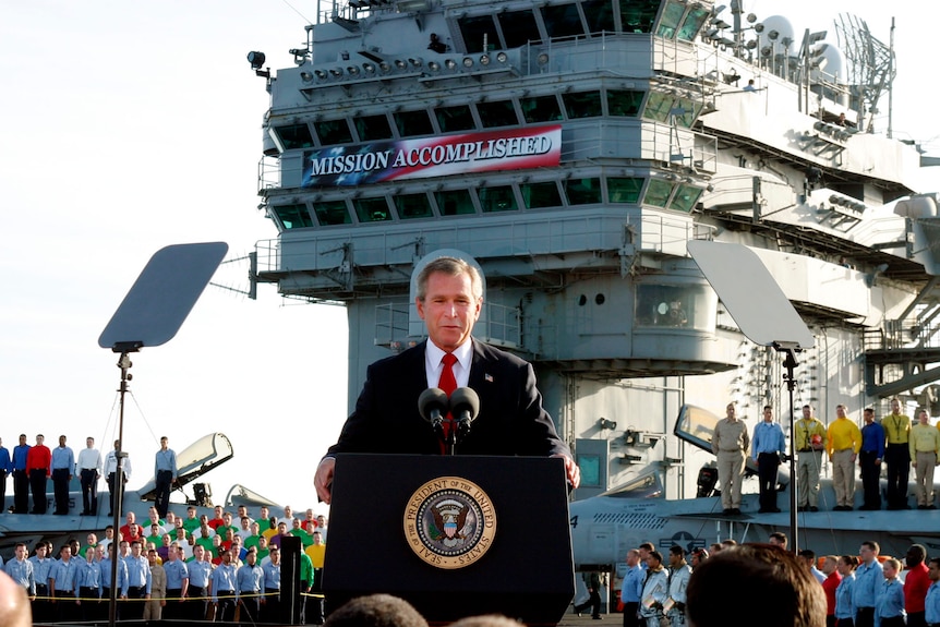 Former US president George W Bush stands aboard an aircraft carrier in front of a banner reading 'Mission accomplished'