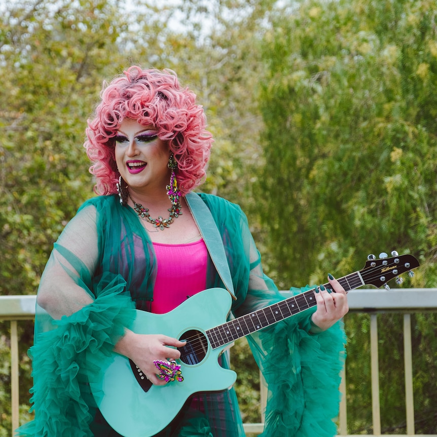 Smiling drag queen holding a guitar. She wears big hair and a flowing dress.