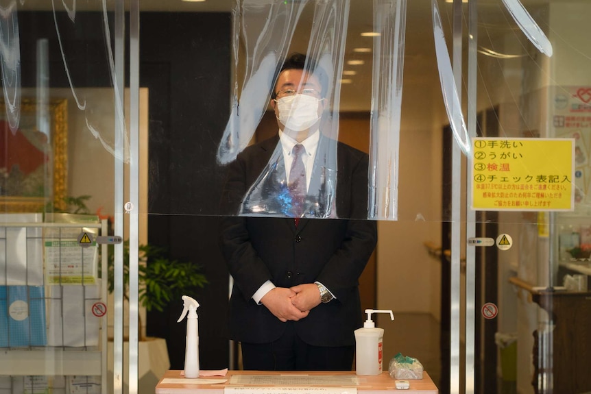 A man in a suit with his hands crossed in front of him and wearing a mask stands behind a screen.