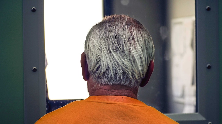 A grey-haired man in a yellow prison jumper stares through a cell window.