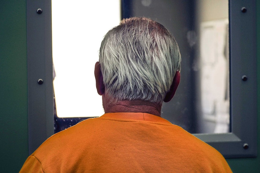A grey-haired man in a yellow prison jumper stares through a cell window.