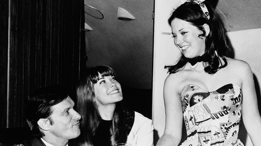 A black and white photo of a young Hugh Hefner and his girlfriend Barbi Benton smiling while they are served a drink.