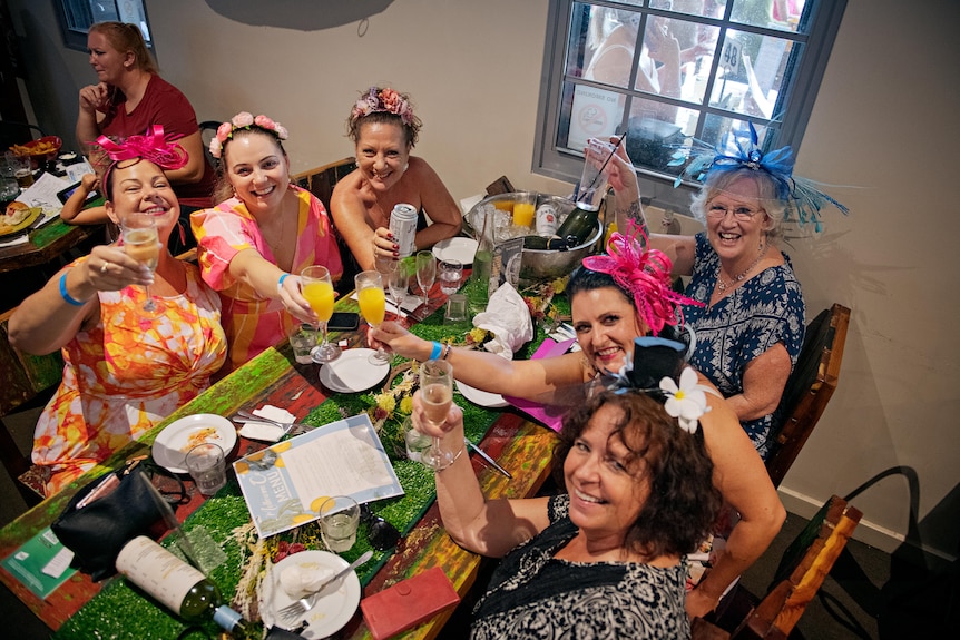 A group of six ladies, all wearing fascinators, cheering their glasses at a pub