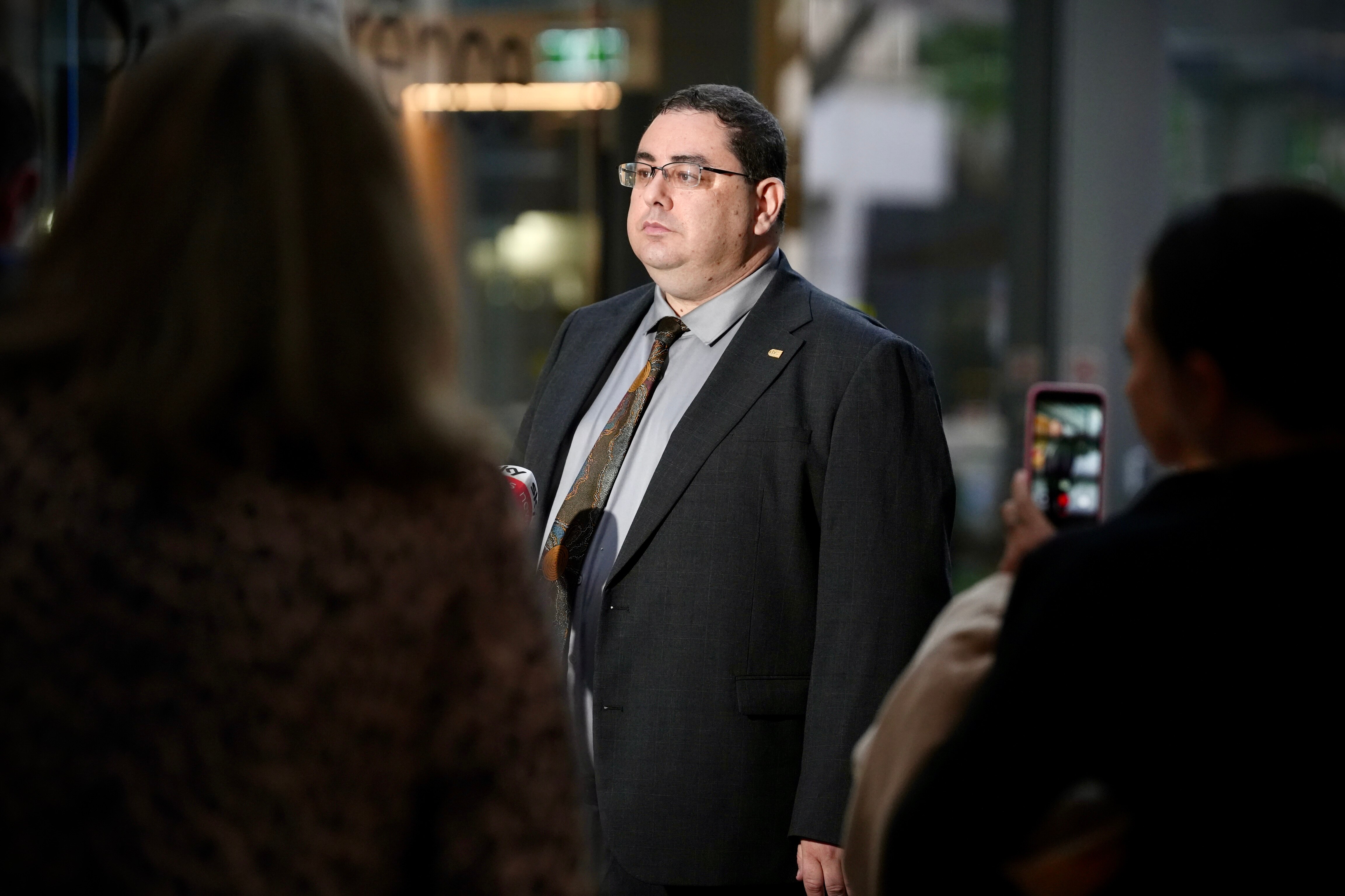 Andre Oboler in a suit, tie, glasses, speaking to a crowd outside an office building.