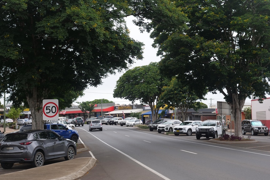 A road lined with trees with several cars driving away and carparks full on either side of the street.