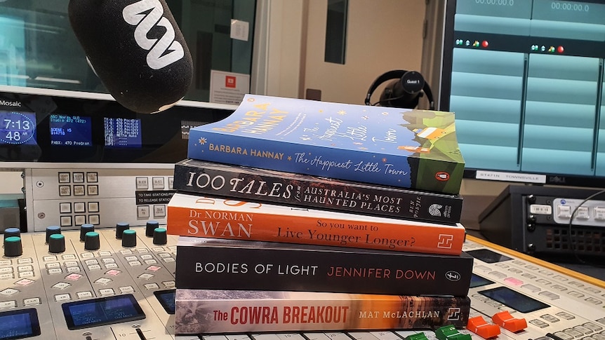 five books stacked on top of a radio mixing desk in between the faders with a microphone and computer in the background
