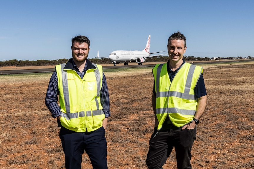 Two men next to a runway wearing high-vis as a plane takes off in the background.  