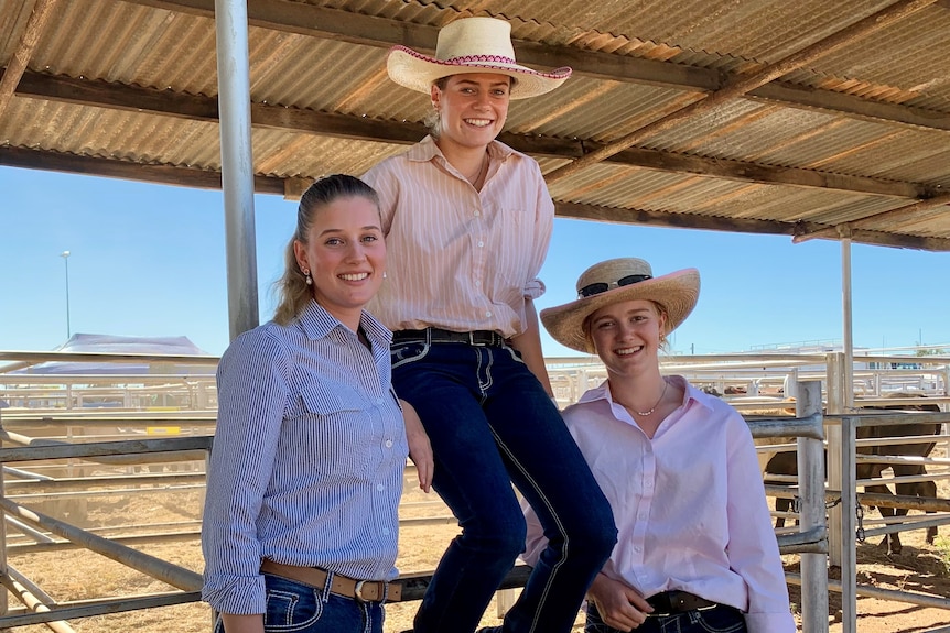 Three young girls at a cattle yard all smiling at the camera, with one sitting on the fence