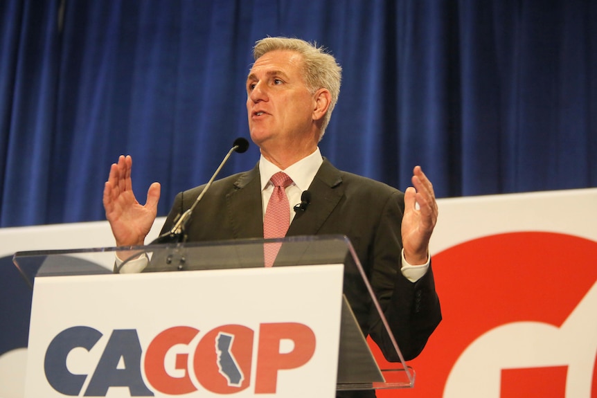 A man is pictured holding his hands up as he speeches, a sign in front of him says 'CAGOP'. 