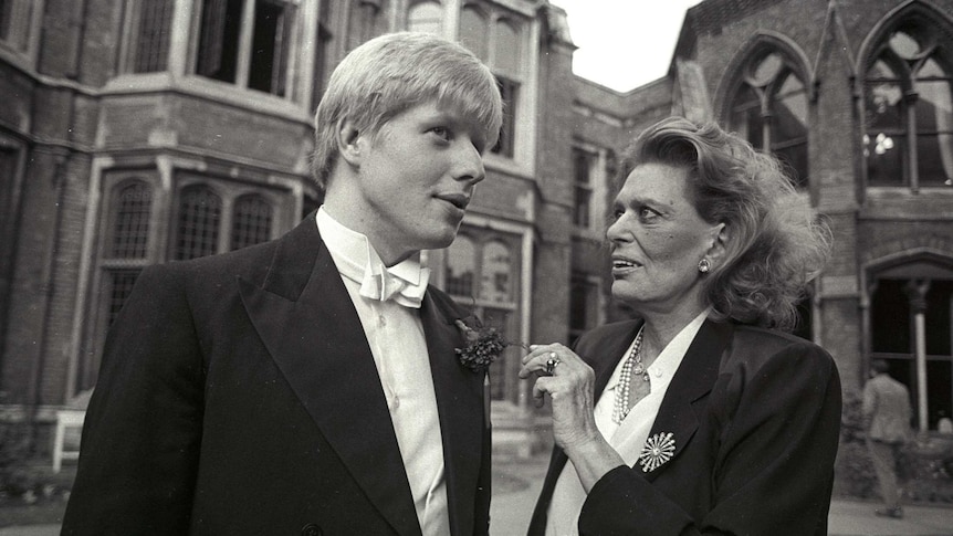 A young Boris Johnson wearing a tuxedo, speaks with a woman wearing a black blazer outside old buildings.