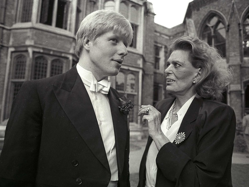 A young Boris Johnson wearing a tuxedo, speaks with a woman wearing a black blazer outside old buildings.
