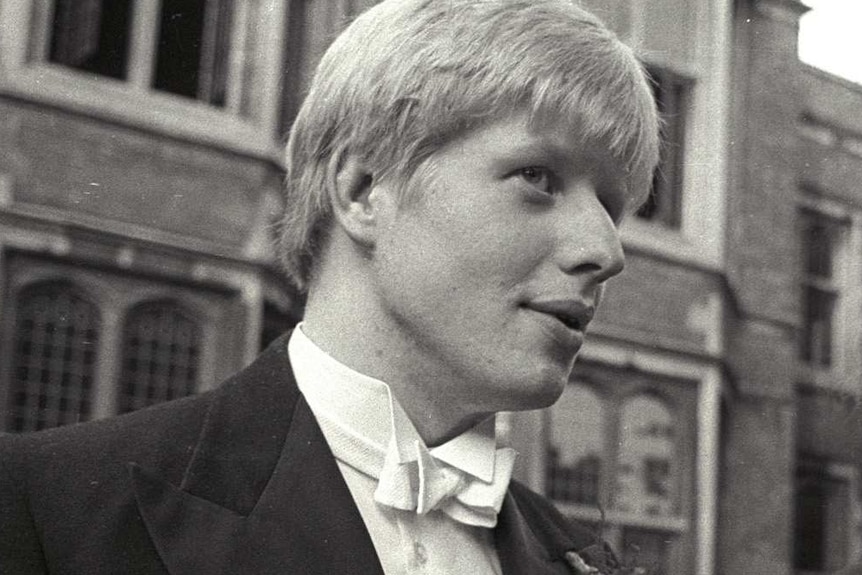 A young Boris Johnson wearing a tuxedo, speaks with a woman wearing a black blazer outside old buildings.