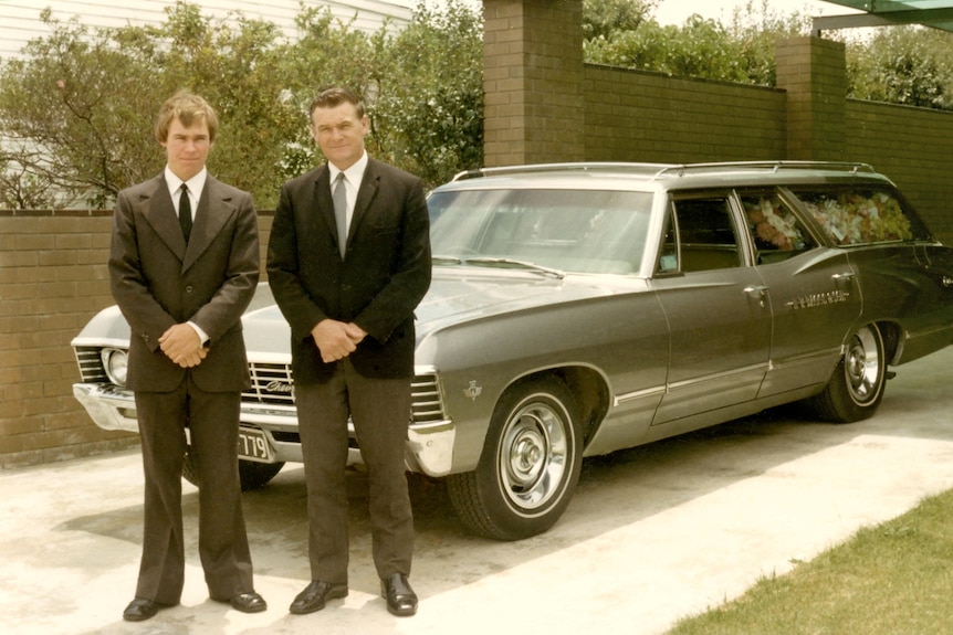 Vintage photo of father and son in suits standing in front of a 1970s hearse.