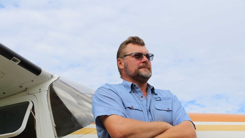 A man stands next to a small plane. he is wearing a blue shirt, sunglasses and has a beard.