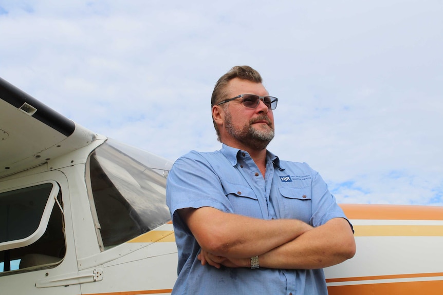 A man stands next to a small plane. he is wearing a blue shirt, sunglasses and has a beard.