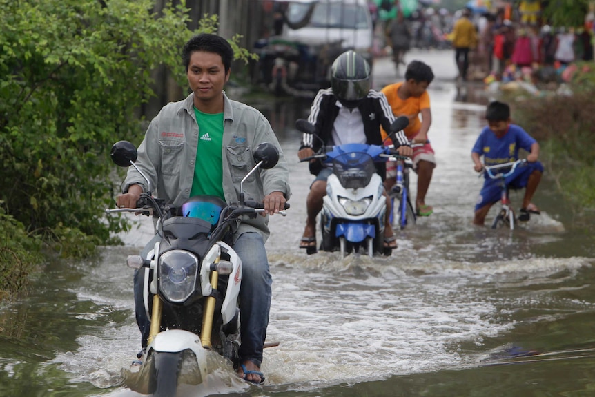 Several people ride motor scooters along a flooded road.