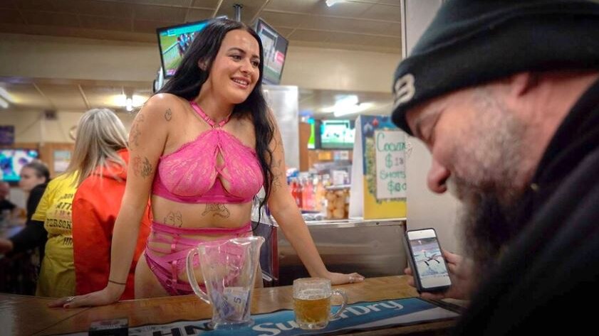 A woman in a hot pink bra working behind a bar, smiling.