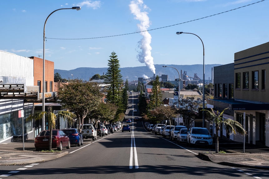 A street in Port Kembla shows houses and cars lining the street, smoke plumes of industrial area billowing in the background