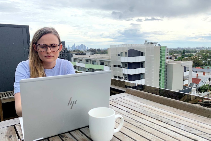 Brighid works on a laptop on the balcony of her apartment. The CBD skyline can be seen in the background.