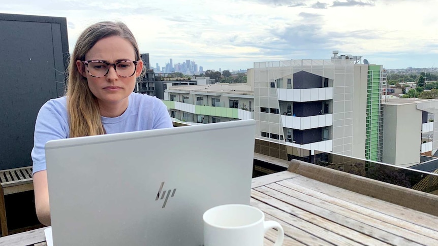 Brighid works on a laptop on the balcony of her apartment. The CBD skyline can be seen in the background.