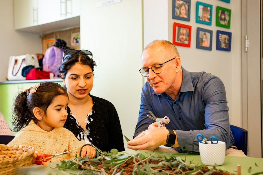 Weatherill sits with a little girl and her mother at a desk covered in plant matter inside a child care facility.